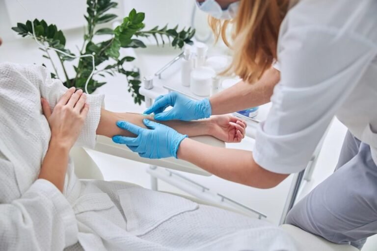 A healthcare professional wearing gloves and a face mask inserts a needle into a seated patient's forearm in a clean clinical setting, likely for a blood draw or IV placement.